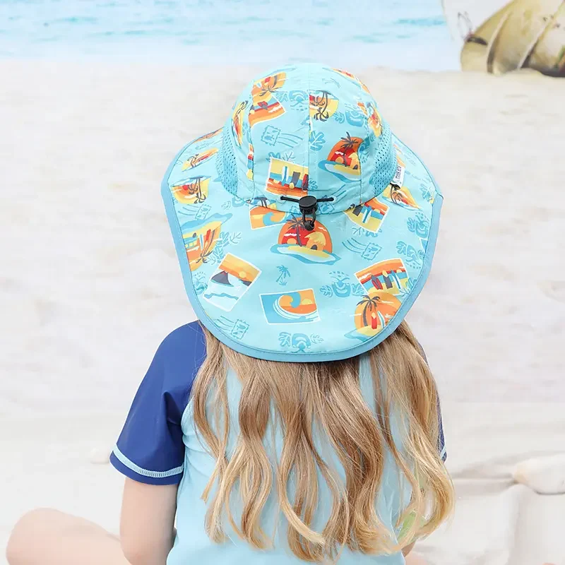 A child with long blonde hair is wearing a light blue sun hat with colorful beach-themed prints. The child is dressed in a light blue and navy shirt and is sitting on a sandy beach, with the ocean in the background. Keywords: Kid's hat, light blue sun hat, beach-themed hat, children's headwear, summer accessories, sun protection, colorful prints, beach fashion, outdoor wear for kids, stylish kid's hat.