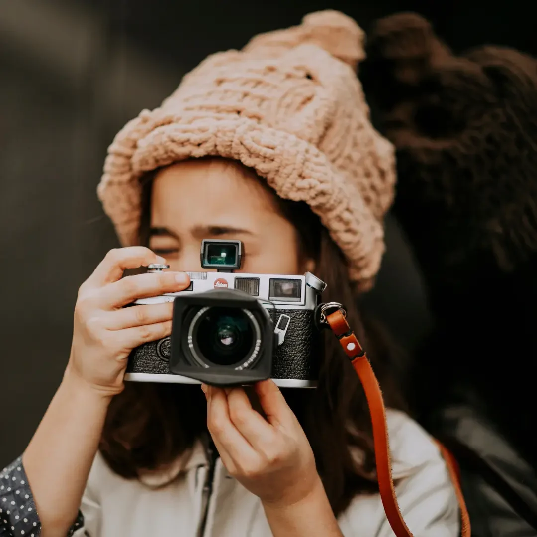 A woman wearing a chunky knitted beige hat is taking a photo with a vintage camera. She is focused on the camera viewfinder, and the background is blurred, emphasizing her and the hat. The scene conveys a cozy and artistic atmosphere. Keywords: Knitted hat, chunky knit hat, beige knit hat, women's headwear, cozy fashion, winter accessories, photography, vintage camera, stylish hat, warm hat, artistic look.