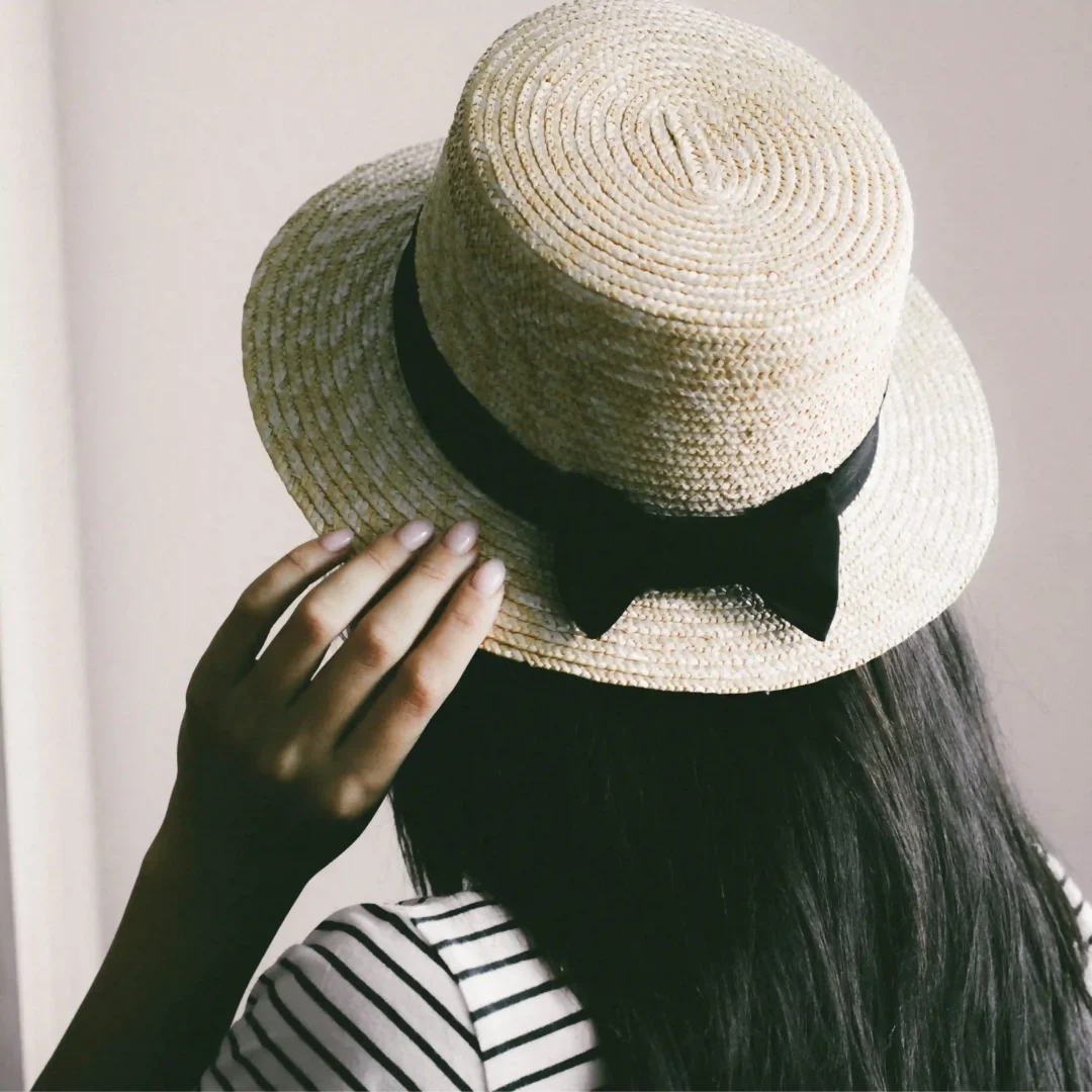 A person with long dark hair is wearing a straw hat with a black ribbon and bow. The individual is dressed in a striped shirt and is shown from behind, touching the brim of the hat. The background is neutral, highlighting the hat's texture and design. Keywords: Straw hat, summer hat, black ribbon hat, boater hat, casual headwear, beach accessories, sun protection, fashion hat, women's straw hat, vintage style, stylish headgear.