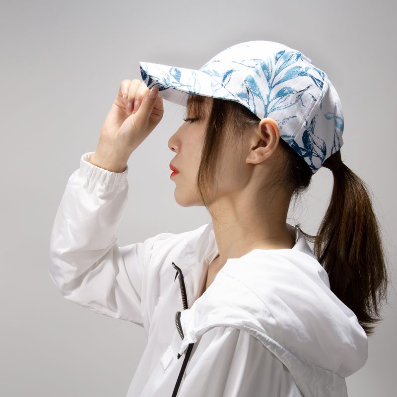 A woman with a ponytail is wearing a white baseball cap with a blue leafy pattern. She is dressed in a white windbreaker jacket and is adjusting the brim of the cap. The background is a plain light gray, highlighting the cap's design. Keywords: Baseball cap, white baseball cap, blue leafy pattern, stylish headwear, casual fashion, women's accessories, outdoor wear, sporty look, trendy cap, fashion statement, adjustable cap.