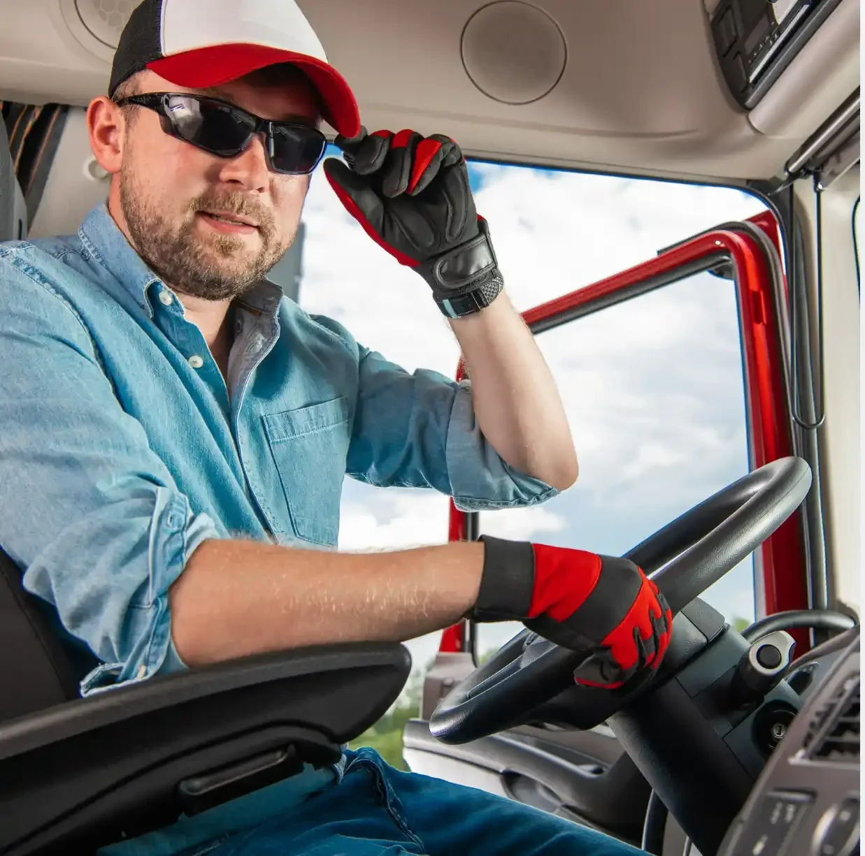 A man wearing a red and white trucker cap and black sunglasses is sitting in the driver's seat of a truck. He is dressed in a light blue denim shirt and wearing black and red gloves, adjusting his sunglasses while looking at the camera. The background shows the interior of the truck cab and a partially visible cloudy sky outside the window. Keywords: Trucker cap, red and white trucker hat, truck driver, men's headwear, casual cap, driving accessories, outdoor workwear, truck cabin, stylish cap, protective gloves, sunglasses.