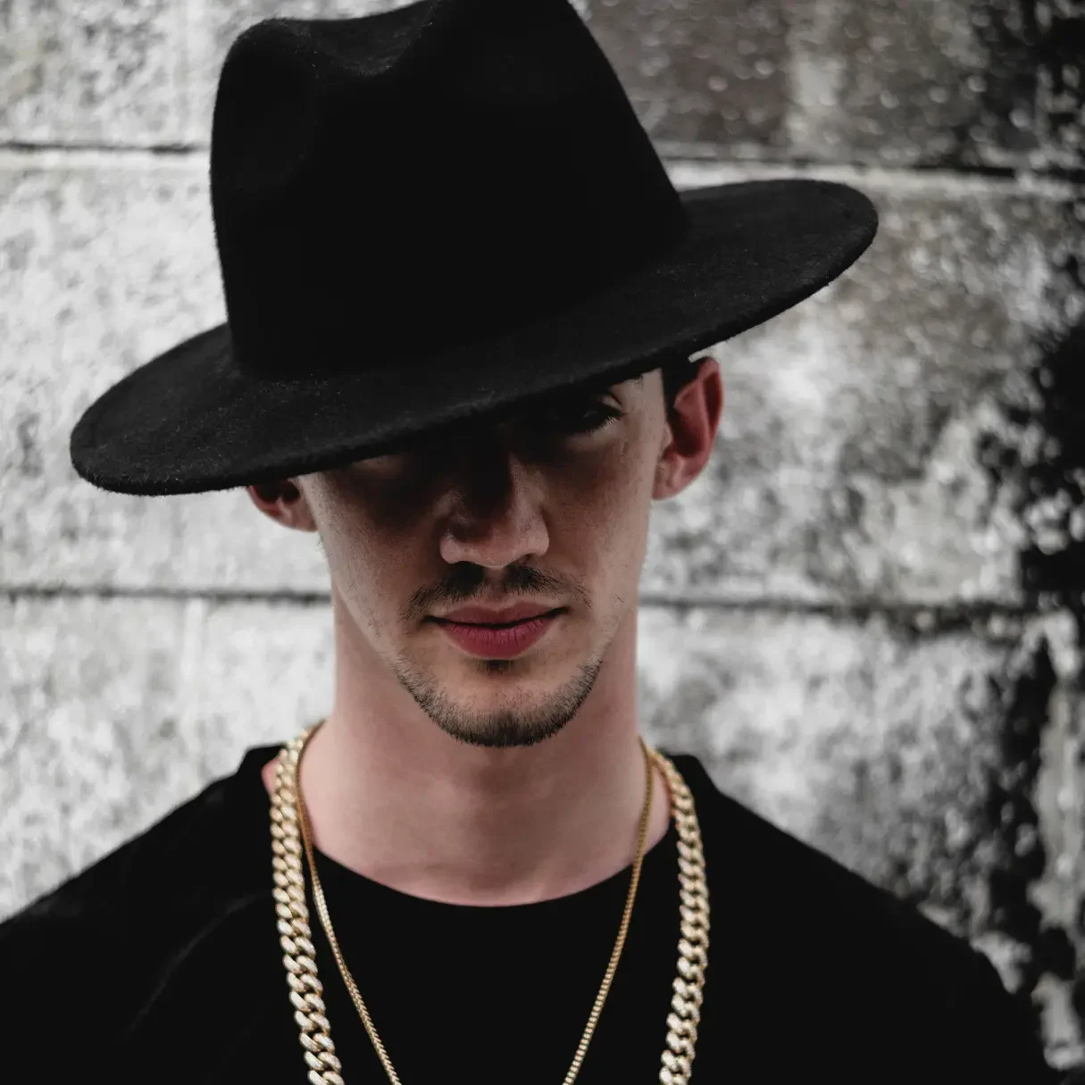 A man wearing a black Panama hat with a wide brim is looking directly at the camera. He is dressed in a black shirt and accessorized with a gold chain necklace. The background features a textured gray wall, adding to the stylish and mysterious look. Keywords: Panama hat, black Panama hat, men's headwear, wide-brim hat, stylish hat, fashion accessory, gold chain, trendy look, men's fashion, classic hat, mysterious style.