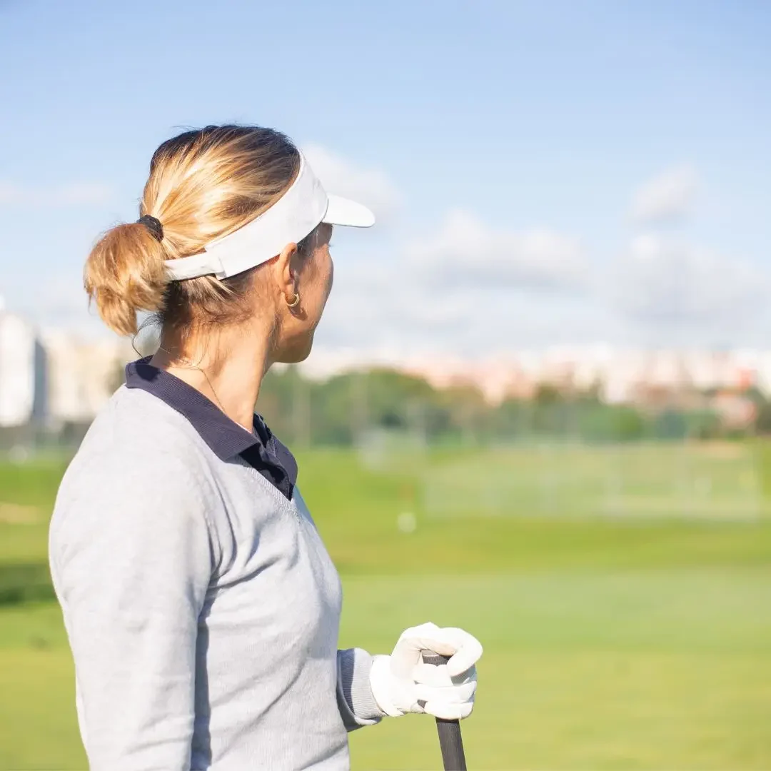 A woman with blonde hair tied in a ponytail is wearing a white sun visor cap while standing on a golf course. She is dressed in a light gray sweater over a dark collared shirt and is holding a golf club. The background features a lush green golf course under a clear blue sky. Keywords: Sun visor cap, white sun visor, golf accessory, headwear, outdoor sports, women's golf fashion, athletic wear, golf course, sporty look, sun protection, casual outdoor attire.