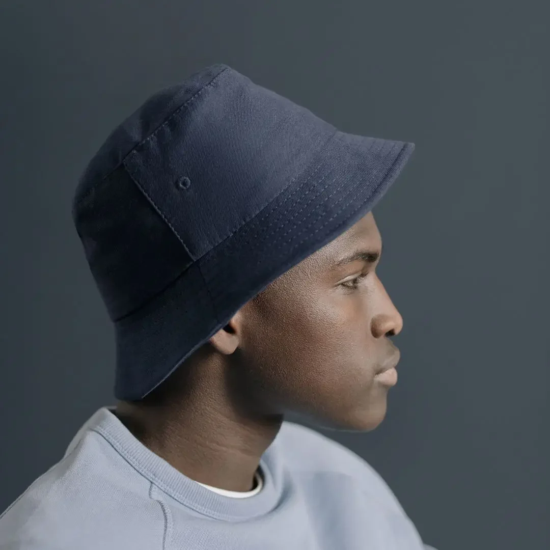 A side profile view of a person wearing a dark blue bucket hat. The individual is dressed in a light gray sweatshirt, and the background is a solid dark gray, emphasizing the hat's design. Keywords: Bucket hat, dark blue bucket hat, fashion accessory, headwear, stylish hat, casual wear, unisex hat, trendy bucket hat, outdoor fashion, side profile.